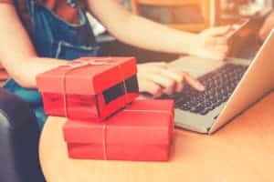 A woman is sitting at a table with red gift boxes and a laptop, carefully managing her financial planning for her kids.