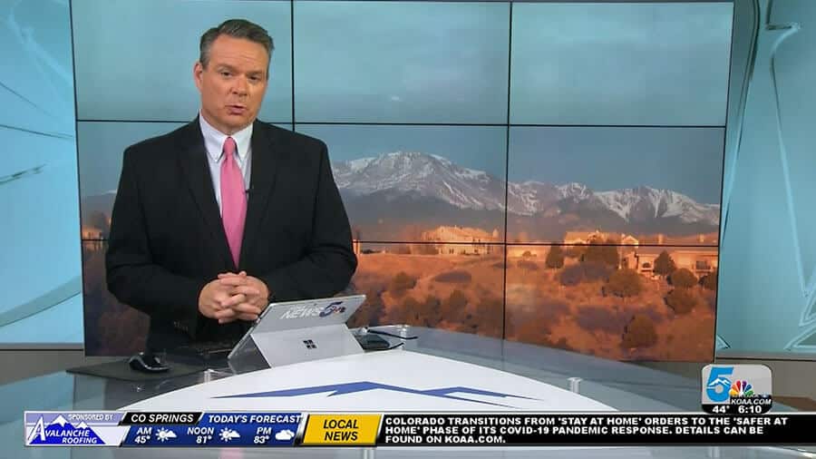 A man in a suit and tie is standing in front of a television screen broadcasting news from KOAA, Colorado Springs-Pueblo's leading channel.