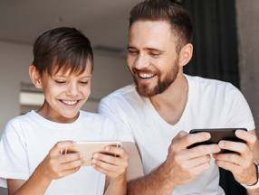 A man and his son are sitting on a couch, both engrossed in their phones. The overwhelmed parent seems buried in work, perhaps checking emails or managing tasks on the BusyKid app.
